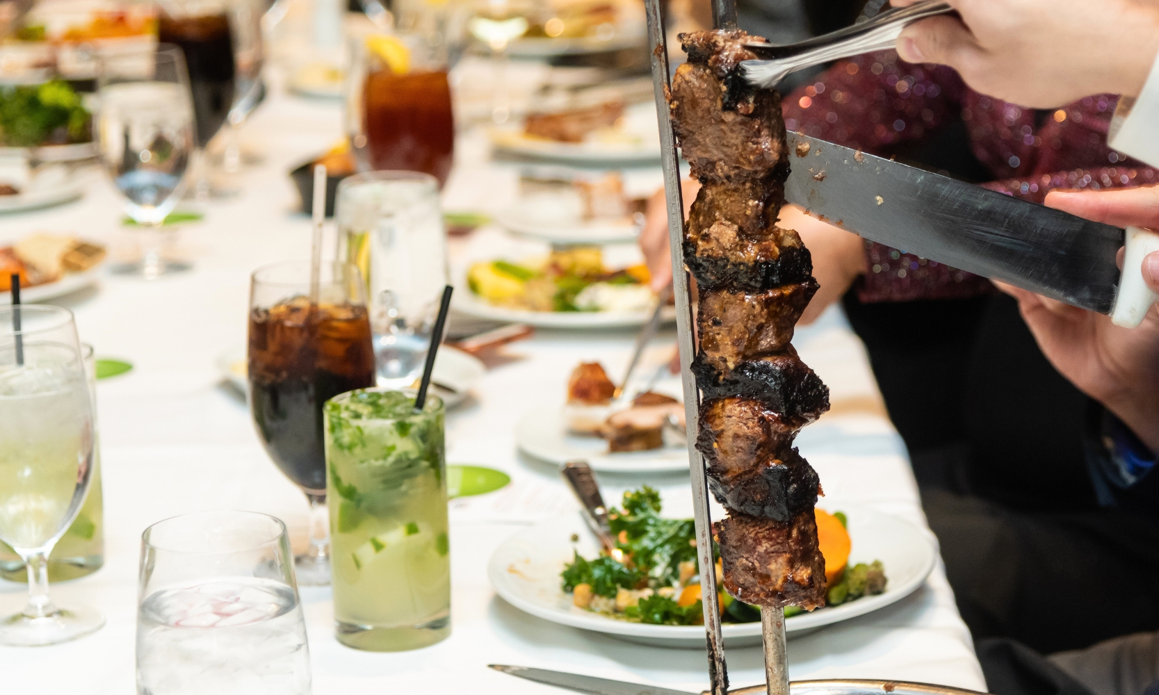 A closeup image of a person cutting a steak skewer for people with drinks and dishes