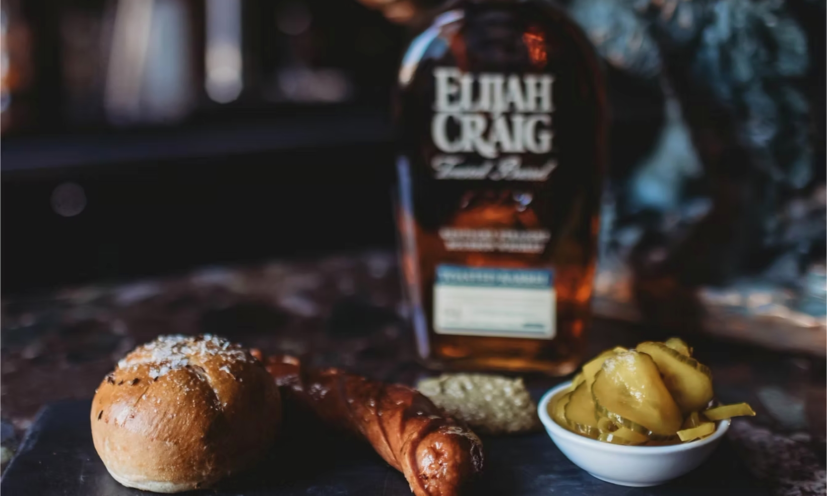 Closeup image of a roll, sausage, and pickles appetizer with a bottle of whiskey at Niche in Geneva, IL