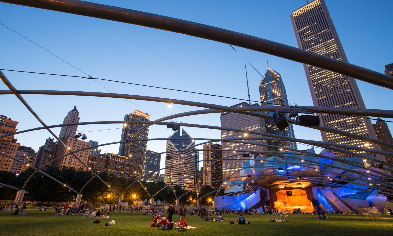 An image of Jay Pritzker Pavillion and The Great Lawn in Millennium Park, Chicago, at night with people sitting