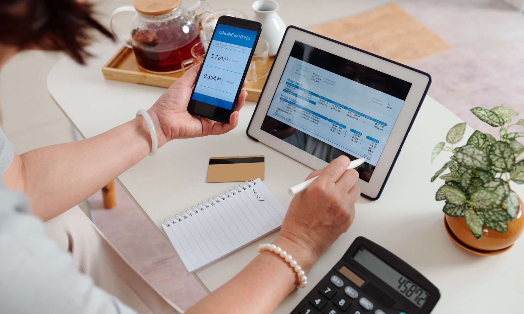 Closeup image of a person calculating their average utilities costs on a tablet and mobile phone with a calculator at a desk