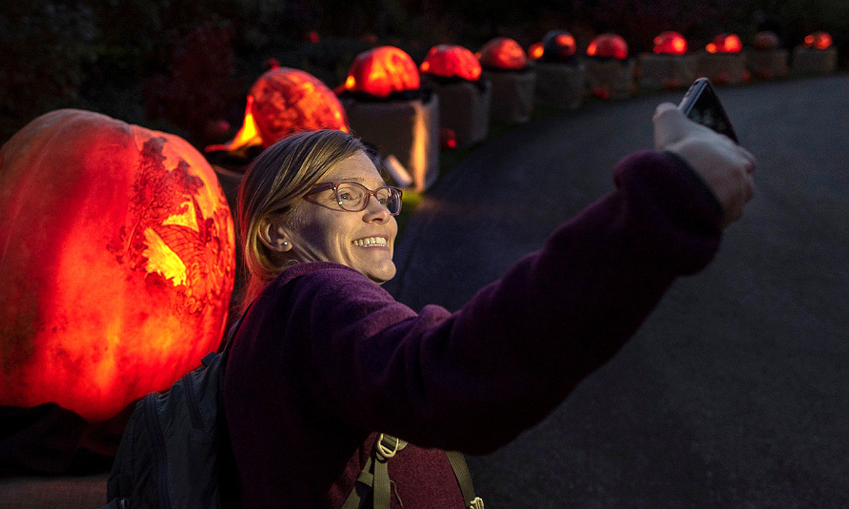 Closeup image of a blonde woman with purple glasses and a sweater in front of lined pumpkins at the Night of 1,000 Jack-o’-Lanterns