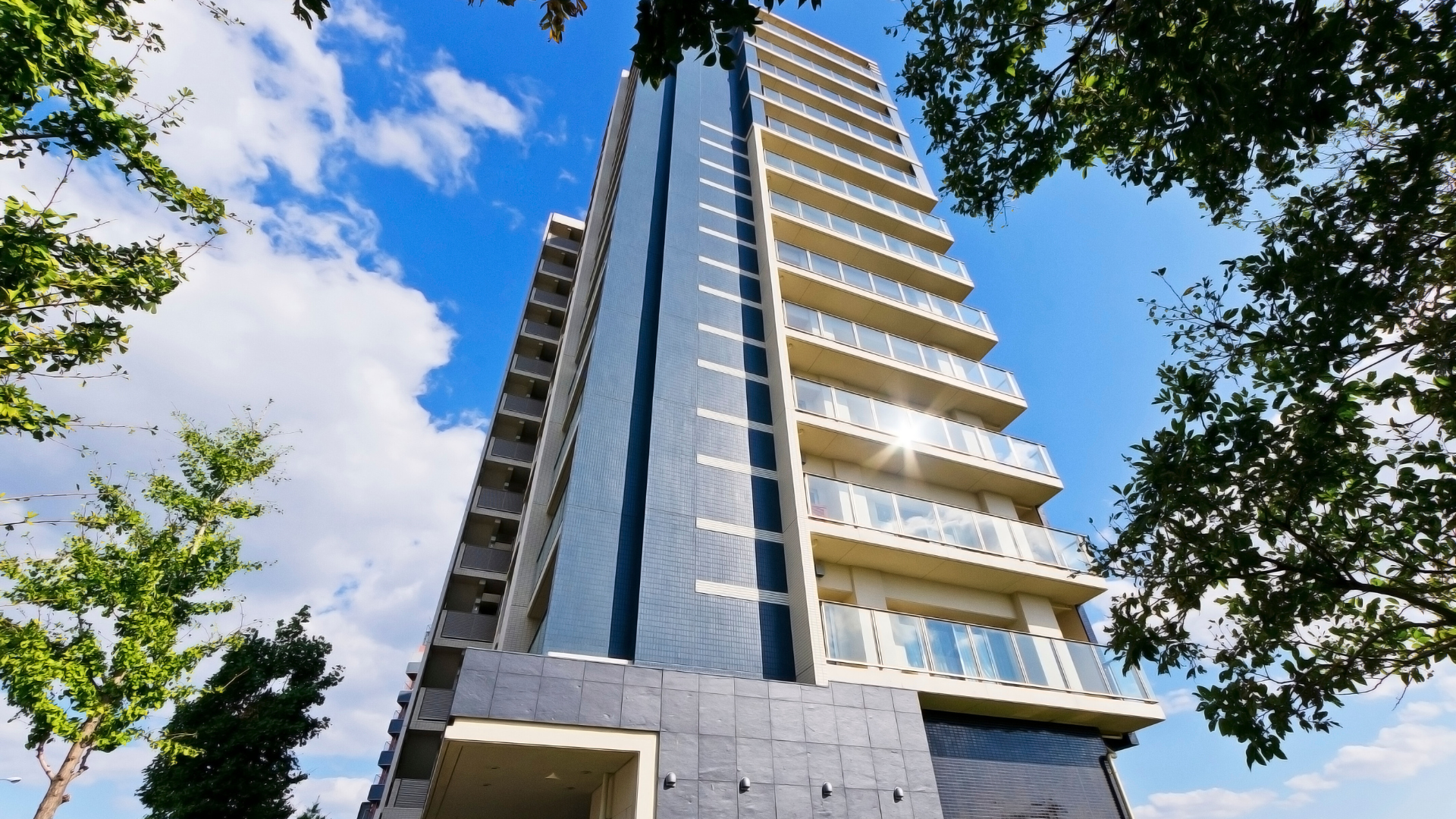 Ground view image of a high-rise apartment building on a partly cloudy day with surrounding trees